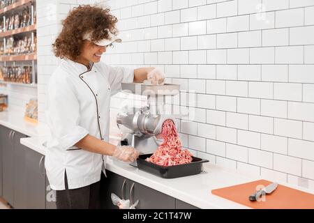 Jeune femme souriante avec des cheveux bouclés mettant de petits morceaux dans le moulin à viande et les grids dans la viande hachée. Vue de la jolie femme travaillant dans la boucherie, préparant la viande pour la vente en magasin. Banque D'Images