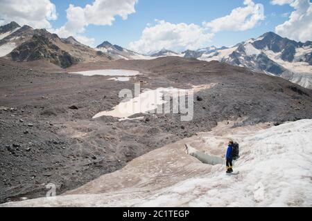 Un alpiniste avec un sac à dos marche dans des crampons le long d'un glacier poussiéreux avec des trottoirs dans les mains entre les fissures dans le Banque D'Images