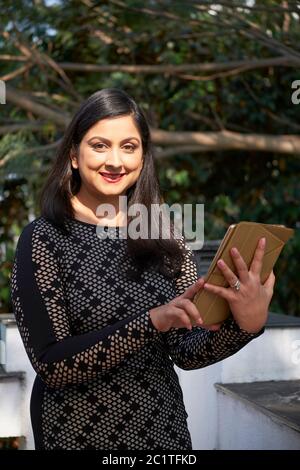 Femme indienne moderne avec de longs cheveux debout sur la terrasse tenant une tablette numérique regardant l'appareil photo sourire, vertical moyen coup Banque D'Images