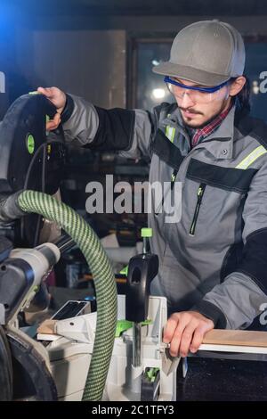 Jeune homme brunette portant une casquette dans une veste grise de profession un charpentier coupe des planches de bois avec une scie circulaire sur un travail Banque D'Images