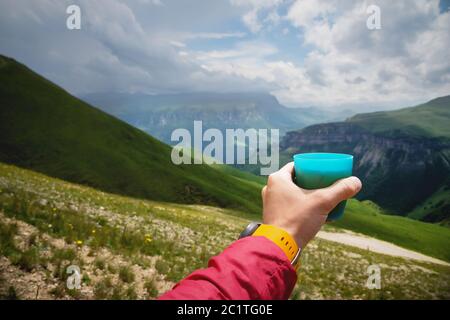 Vue de première personne d'une main d'homme tenant une tasse de thé en plastique contre un plateau de collines vertes et un ciel nuageux en été Banque D'Images