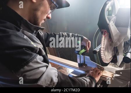 Jeune homme brunette portant une casquette dans une veste grise de profession un charpentier coupe des planches de bois avec une scie circulaire sur un travail Banque D'Images