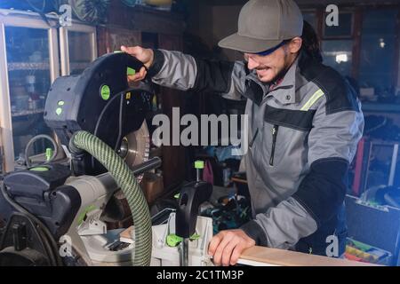 Jeune homme brunette portant une casquette dans une veste grise de profession un charpentier coupe des planches de bois avec une scie circulaire sur un travail Banque D'Images