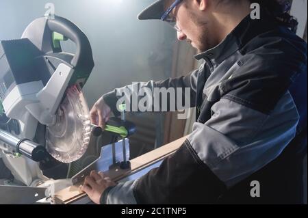 Jeune homme brunette portant une casquette dans une veste grise de profession un charpentier coupe des planches de bois avec une scie circulaire sur un travail Banque D'Images