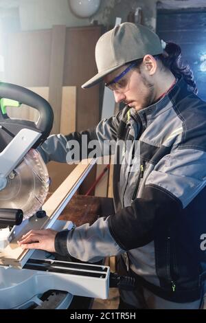 Jeune homme brunette portant une casquette dans une veste grise de profession un charpentier coupe des planches de bois avec une scie circulaire sur un travail Banque D'Images