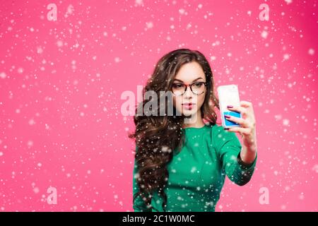Jolie fille brune avec de longs cheveux ondulés montrant bâton dehors et prenant autoportrait sur son smartphone. Porter une veste verte et Banque D'Images
