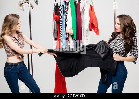 Récolte de deux jeunes émotionnels et stylés femme tirant des vêtements et arguant pour elle. Action dans la cabine d'essayage contre le rack de caillot Banque D'Images