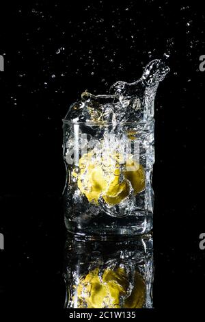 Un verre d'eau placé sur une surface réfléchissante avec un fond noir et de l'eau éclabousse après avoir déposé un citron dans le verre. Concept été Banque D'Images