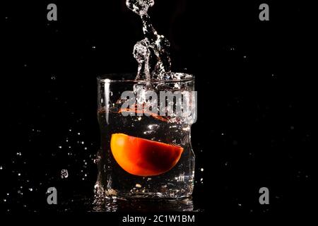 Un verre d'eau dans un fond sombre et l'eau étant éclaboussé avec un morceau de tomate coupée tombée à l'intérieur du verre Banque D'Images