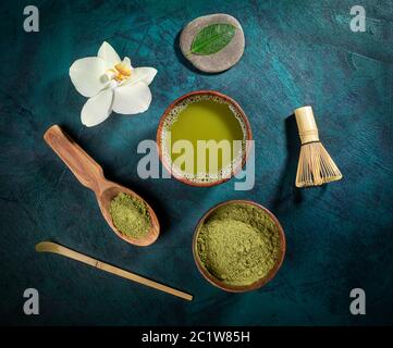 Set pour la cuisson du thé matcha: Tasses en bois de thé et de poudre, fouet, cuillères, feuille et fleur d'orchidée disposés sur fond émeraude. Le thé Matcha est sain b Banque D'Images