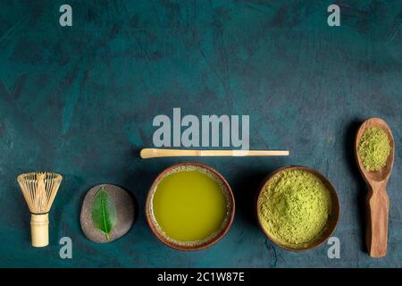 Set pour la cuisson du thé matcha: Tasses en bois avec thé et poudre, fouet, cuillère avec poudre, feuilles disposées en rangée sur fond émeraude. Le thé Matcha est riche Banque D'Images