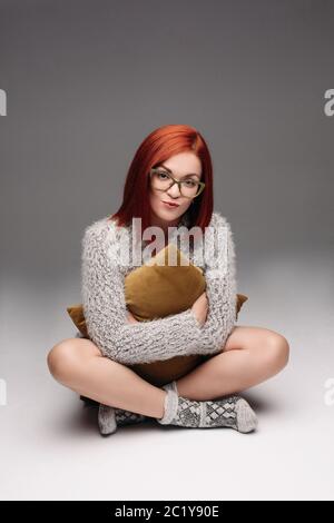 Fille aux cheveux rouges dans chandail gris et des chaussettes chaudes assis sur le plancher dans le style turc et tristement looking at camera. Banque D'Images