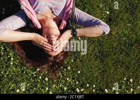 Jeune femme souriante, couchée sur l'herbe avec de petites fleurs de Marguerite, fermant les yeux avec les mains. Vue de dessus Banque D'Images