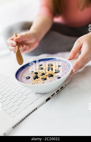 Gros plan sur une femme enceinte qui mange du muesli avec des myrtilles tout en travaillant sur un ordinateur portable. Banque D'Images