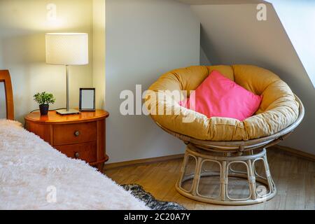 Chaise en bois avec oreillers dans le coin. Chevet avec lampe et fleur. Plafond incliné. Intérieur moderne de la chambre. Banque D'Images