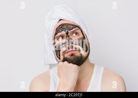 Spa et soins de la peau pour les hommes, hygiène du visage et du corps. Jeune homme avec le visage concentré rêvant, la tête blanche de serviette Oh et le masque noir de charbon de bois sur son visage Banque D'Images