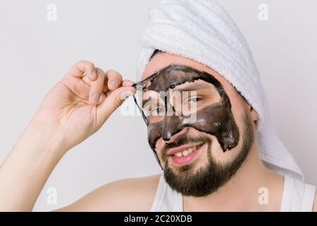 Beau sourire barbu gars est enlever le masque noir purifiant de son visage sur fond blanc. Portrait de l'homme en gros plan pendant le traitement au spa Banque D'Images