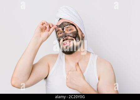 Spa et soins de la peau pour hommes. Jeune drôle gars avec noir anti acné masque de charbon de bois sur son visage sur fond blanc, enlever le masque du visage et de montrer Banque D'Images