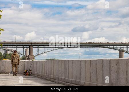 Un homme en costume spécial avec son dos contre un pont automobile et un ciel bleu avec des nuages blancs se tient à côté des cannes à pêche sur le remblai de la ville Banque D'Images