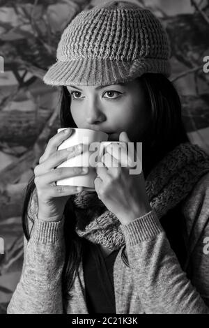 Portrait noir et blanc d'une jeune fille vêtue de vêtements pour le temps froid et buvant d'une tasse blanche. Banque D'Images