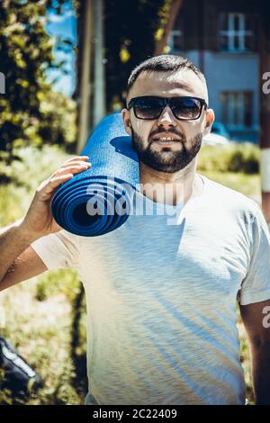 Jeune sportif barbu tenant un tapis bleu au parc. Jeune homme pratiquant le yoga exercice de fitness en plein air. Banque D'Images