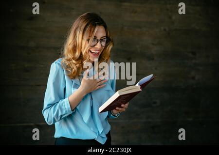 Charmante jeune femme d'éclater de rire après avoir lu quelque chose d'amusant dans le livre. Vêtu d'un élégant chemisier, dans les lunettes. Banque D'Images