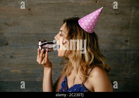 Jolie femme manger un gâteau au chocolat à la crème, la célébration d'anniversaire, de porter un chapeau. Vue de profil. Banque D'Images
