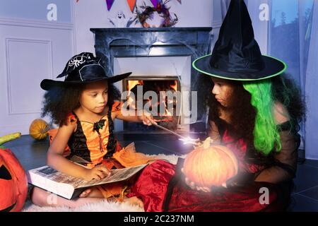 Deux filles en costumes de sorcière Halloween ont jeté un sort sur une citrouille avec baguette magique tenant le livre d'assistant assis près de la cheminée Banque D'Images