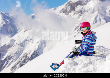 Belle fille en casque rose et équipement de ski s'assoir sur le sommet de la montagne dans la neige observant la vue Banque D'Images