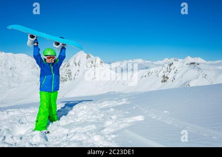 Un adorable garçon se tient dans la neige profonde tenir le snowboard sur les mains levées et porter un casque de ski sur un panorama de montagne propre avec un espace de copie Banque D'Images