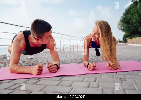 Vue avant du jeune sportif et de la sportive à la recherche de l'autre tout en faisant plank, lors de l'entraînement près de la mer. Sport concept. Banque D'Images