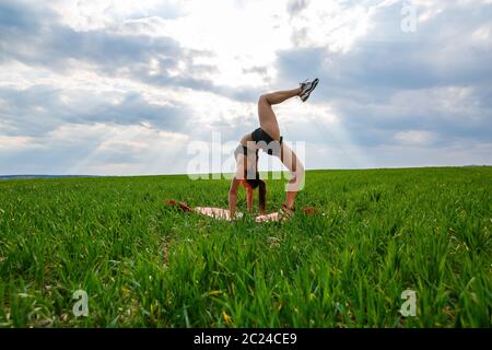 Une jeune femme en haut et short noirs exécute une main. Un modèle se tient sur ses mains, faisant des fractionnements de gymnastique contre le ciel bleu. Un mode de vie sain Banque D'Images