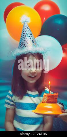 Portrait of a cute little girl wearing hat de fête d'anniversaire et s'apprête à souffler une bougie sur un gâteau d'anniversaire, fête décorée de beaucoup de col Banque D'Images