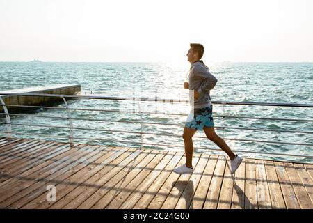 Profil des jeunes sportifs motivés, le jogging sur le quai, près de l'océan. Pleine longueur. Vêtus de shorts de sport et de l'enveloppe. À l'extérieur. Banque D'Images
