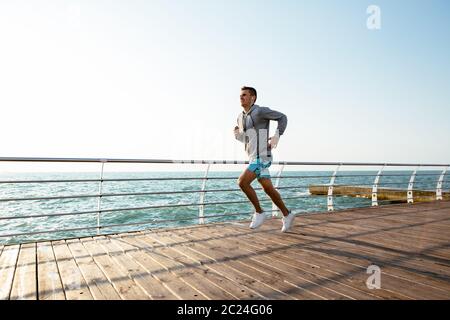 Vue latérale du jeune sportif en cours avec visage fatigué, dans un casque, écouter de la musique. Près de la mer. Vêtus de shorts de sport et de l'enveloppe. Banque D'Images