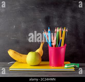 Vert pomme, banane mûre sur une pile de carnets, papeterie rouge verre avec multi-couleur crayons en bois sur l'arrière-plan d'un boa craie noire vide Banque D'Images