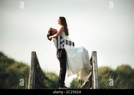 Couple de mariage souriant et heureux embrassant sur un pont en bois à la lumière du soleil du soir. Marié soulevant la mariée à l'air. Copier l'espace. Concept de mariage. Banque D'Images