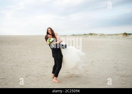 Couple de mariage debout et embrassant à la plage de sable. Marié soulevant la mariée à l'air. Souriant et joyeux, la mariée regarde l'appareil photo. Concept de mariage. Banque D'Images