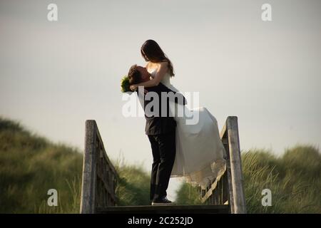 Des jeunes mariés s'embrassant sur un pont en bois dans la campagne en soirée, sous le soleil. Marié soulevant la mariée à l'air. Copier l'espace. Concept de mariage. Banque D'Images