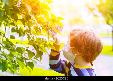 Mignon garçon d'enfant dans le masque médical dans les lilas fleurs nouant à l'odeur des fleurs. Joyeux enfant caucasien à la journée ensoleillée à l'extérieur dans le parc. Allergie et Banque D'Images