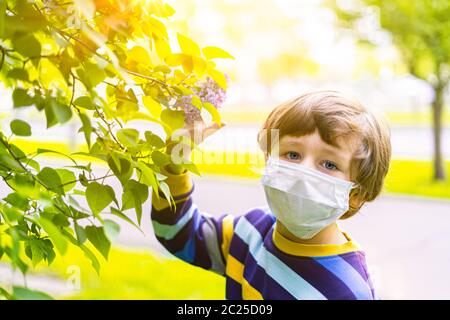 Mignon garçon d'enfant dans le masque médical dans les lilas fleurs nouant à l'odeur des fleurs. Joyeux enfant caucasien à la journée ensoleillée à l'extérieur dans le parc. Allergie et Banque D'Images