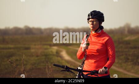 Une femme sportive de race blanche mange une balade en bar à protéines sur le VTT sur la nature. Jeune athlète sportive dans le casque de repos pendant que bi Banque D'Images