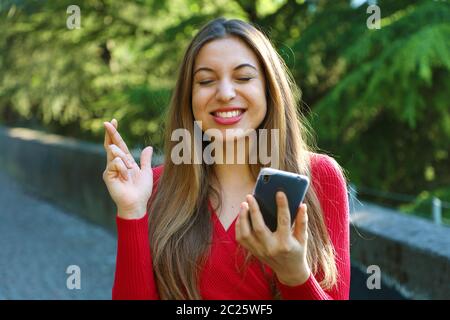 Fille d'espoir crossing fingers holding un smartphone dans l'attente de nouvelles à l'extérieur. Jeune femme avec des doigts de passage et smart phone qui souhaitent le meilleur outsid Banque D'Images