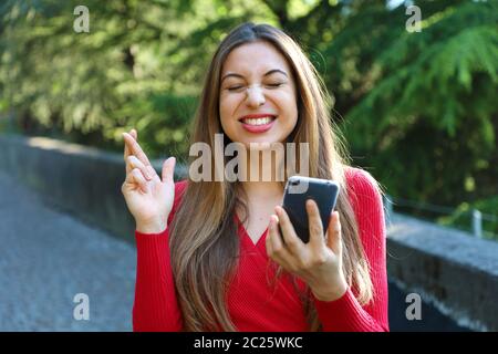 Fille d'espoir crossing fingers holding un smartphone dans l'attente de nouvelles à l'extérieur. Jeune femme avec des doigts de passage et smart phone qui souhaitent le meilleur outsid Banque D'Images