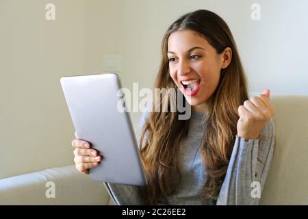 Une femme gagnante excitée tenant un Tablet pc assis sur un canapé à la maison. Joyeuse femme avec une tablette PC regardant son PC à clavier tactile. Banque D'Images