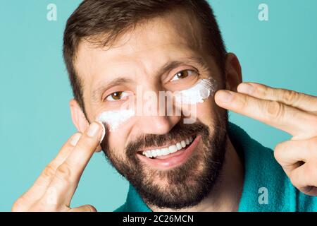 Le sourire joyeux homme beau est application de crème pour le visage des hommes sur son visage sur fond turquoise. Banque D'Images