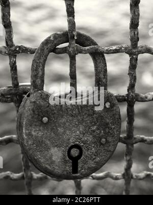 gros plan monochrome foncé d'un ancien cadenas rouillé sur une clôture métallique corrodée sur un arrière-plan extérieur flou Banque D'Images