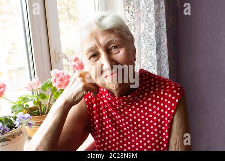 Vieille Femme solitaire assis près de la fenêtre de sa maison avec des fleurs Banque D'Images