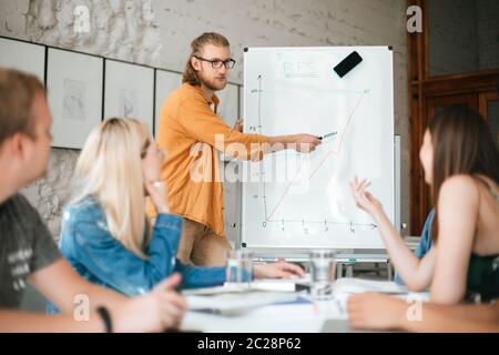 Portrait d'un jeune homme debout près de la planche et parlant avec des collègues au bureau . Groupe de jeunes assis à la table sur la réunion d'affaires et Banque D'Images