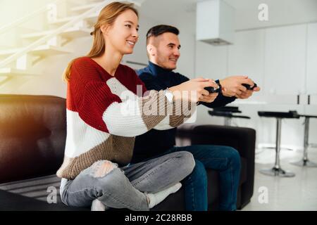 Jeune couple drôle de jouer à des jeux vidéo en utilisant les contrôleurs de jeu, assis sur le canapé à la maison. Habillé dans des vêtements décontractés. Vue de côté. Banque D'Images